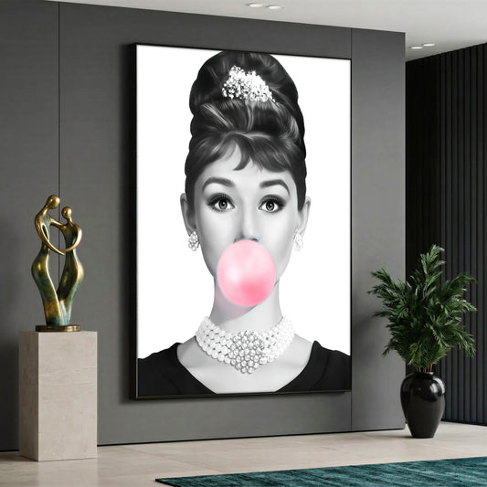 Large vertical black and white canvas print of Audrey Hepburn with a pearl necklace blowing a pink bubble gum, styled in a modern grey interior.